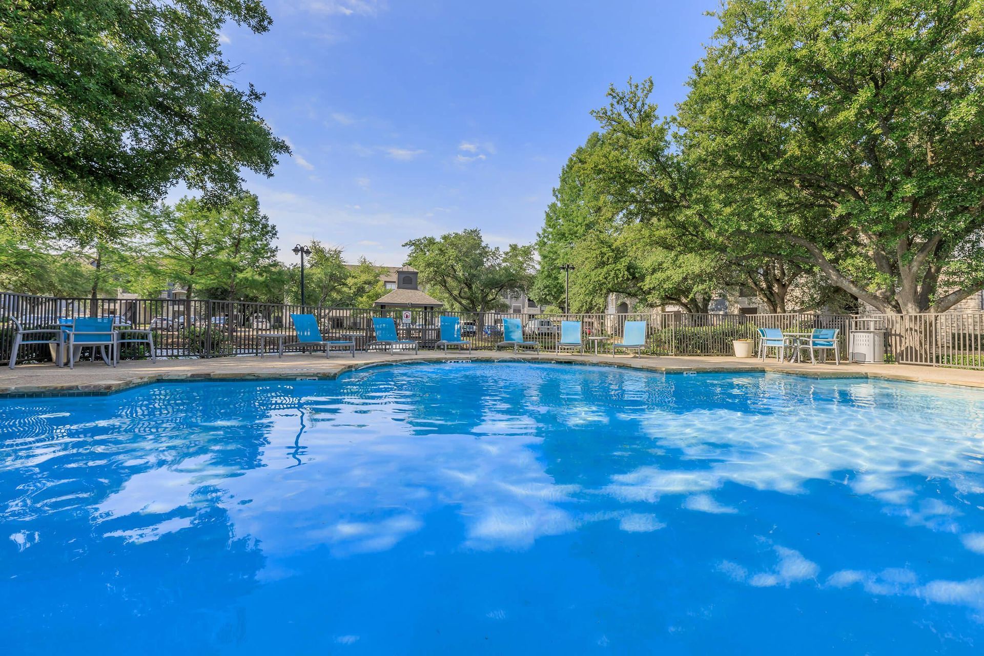 A bright blue swimming pool surrounded by trees and blue lounge chairs on a sunny day.