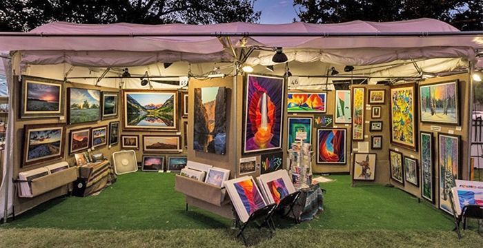 Art booth at an outdoor fair displaying framed paintings in a variety of sizes and vibrant colors. Booth has a white canopy and green turf.