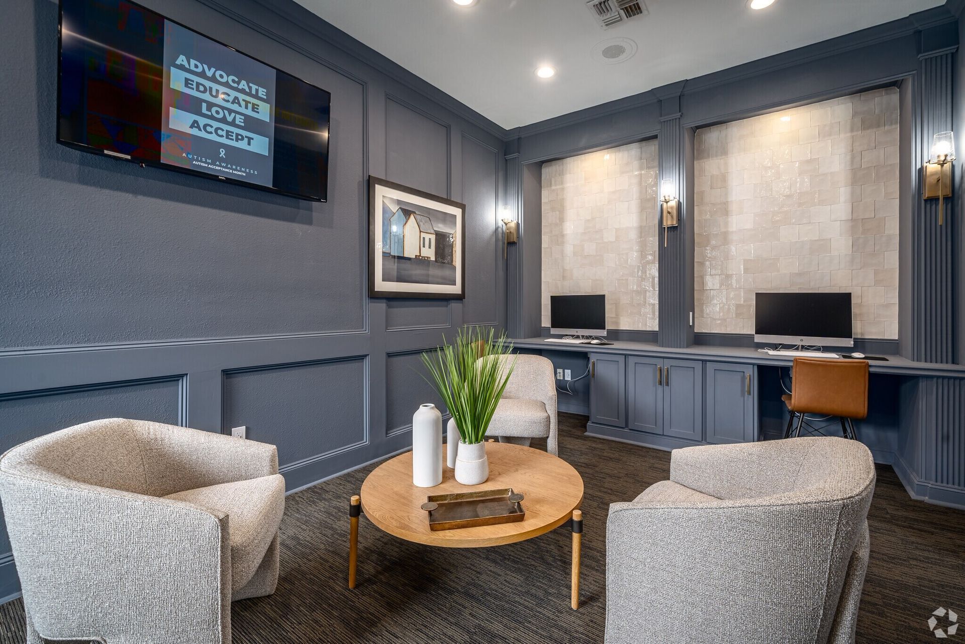 Lounge area with gray walls, two chairs, round coffee table, desks with computers, and wall-mounted art.