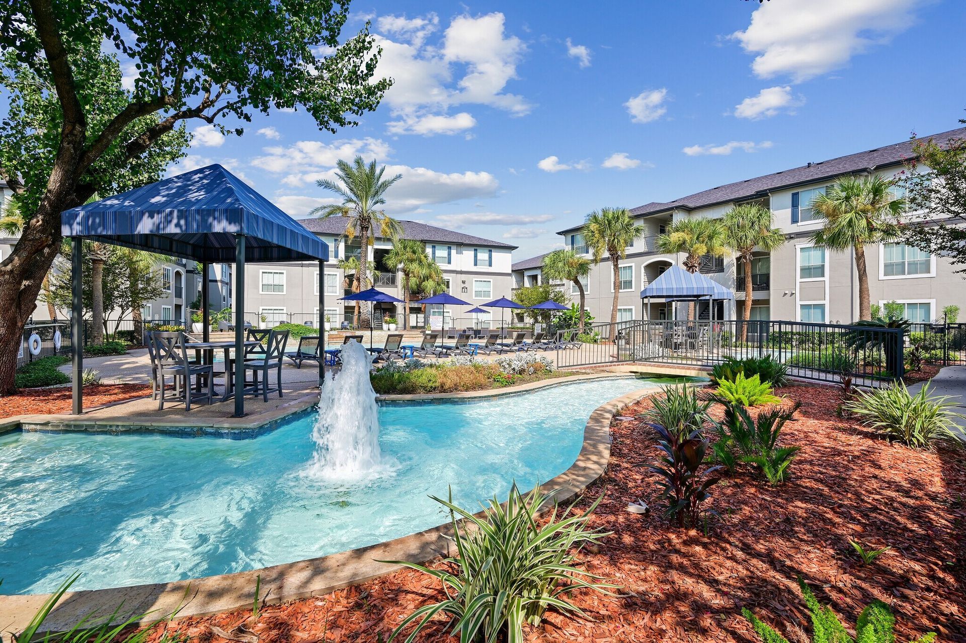 Pool area with water feature, blue canopies, and apartment buildings on a sunny day.