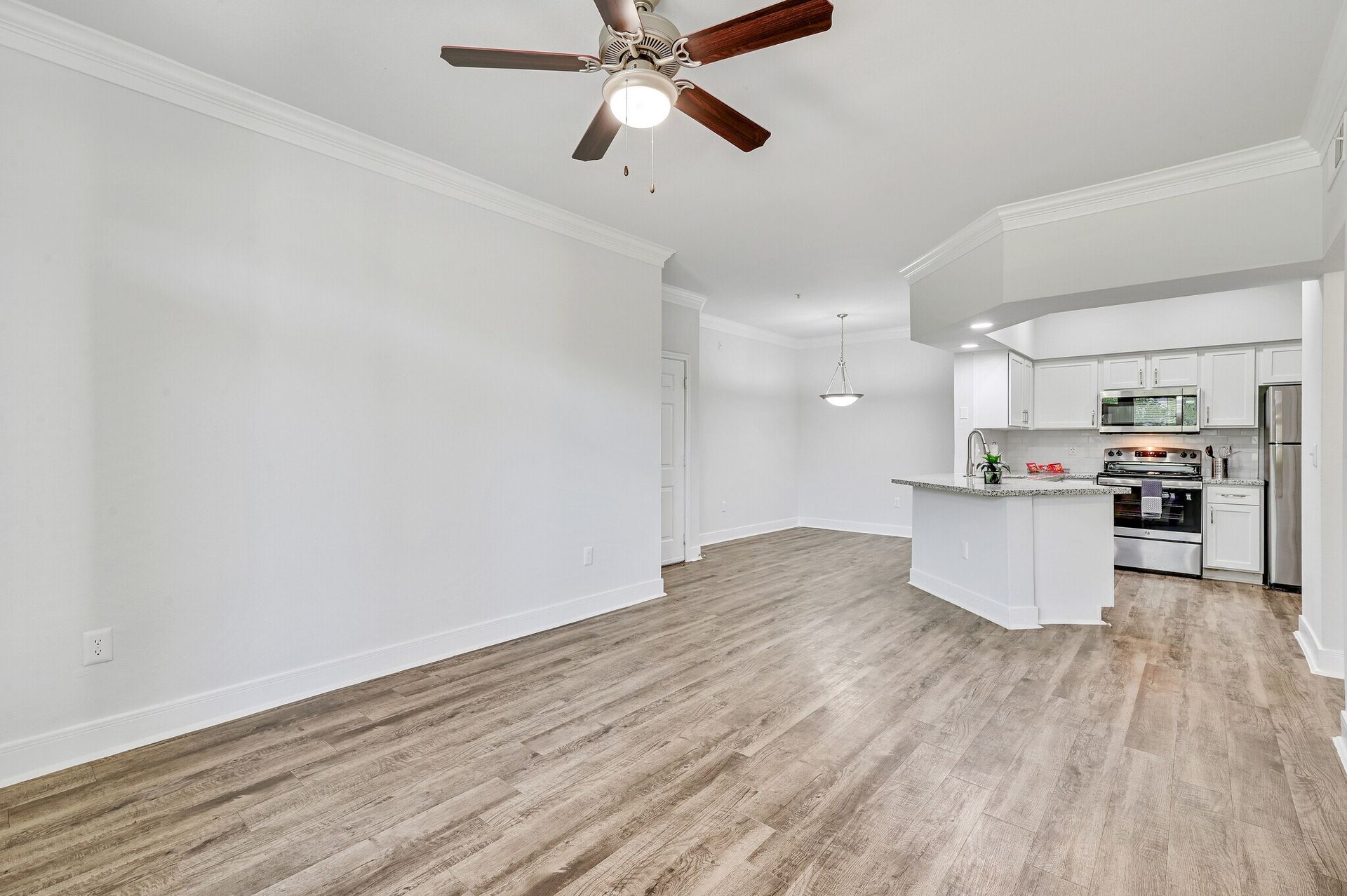 Empty living space with hardwood floors, white walls, and an open kitchen.