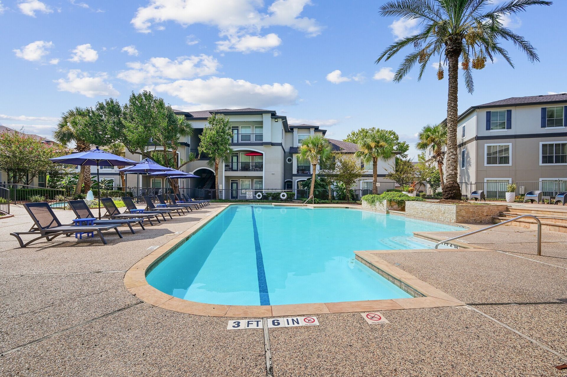 Pool area with lounge chairs, palm trees, and apartment buildings.