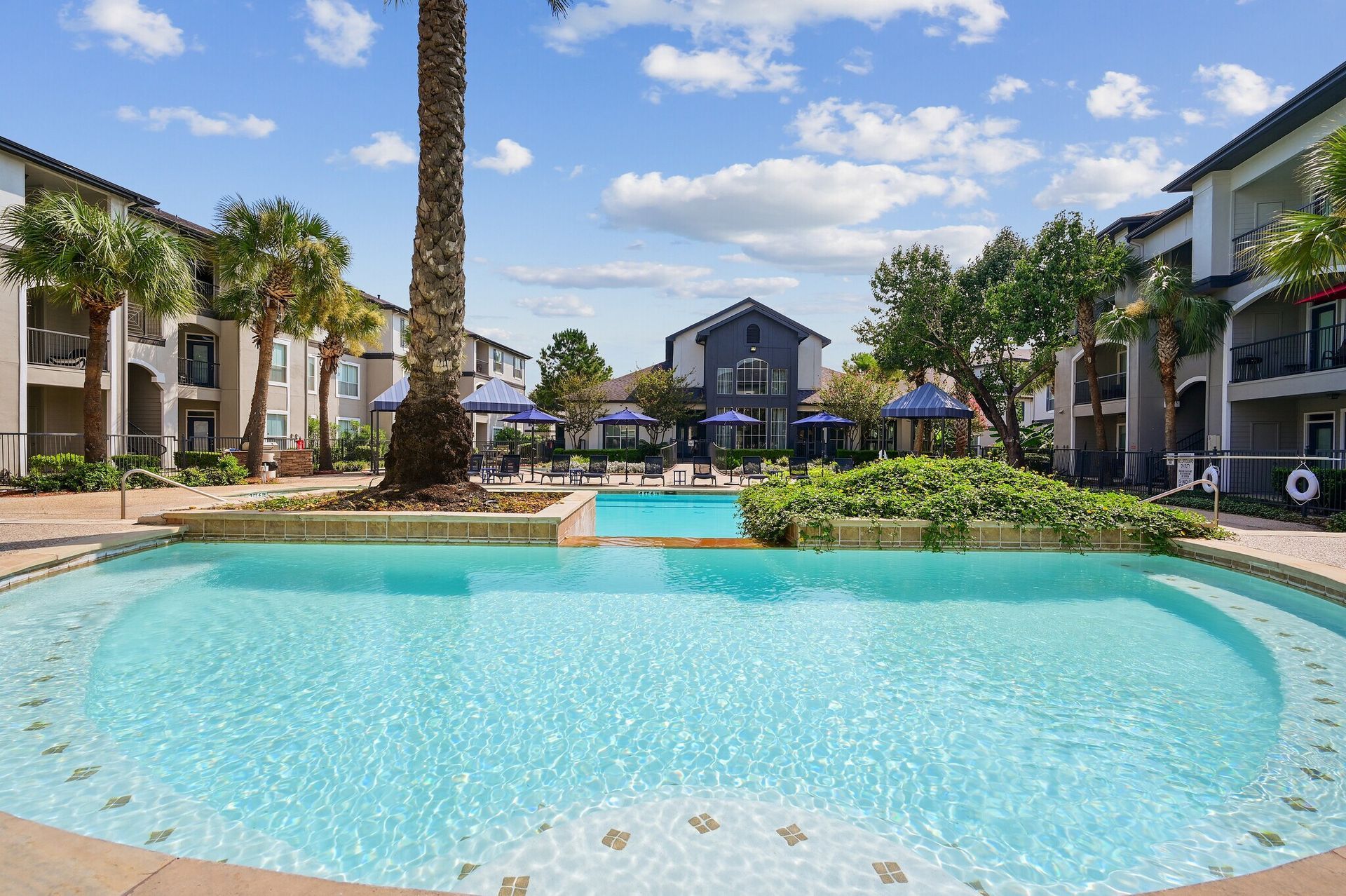 Pool at apartment complex with buildings and palm trees. Sunny day.