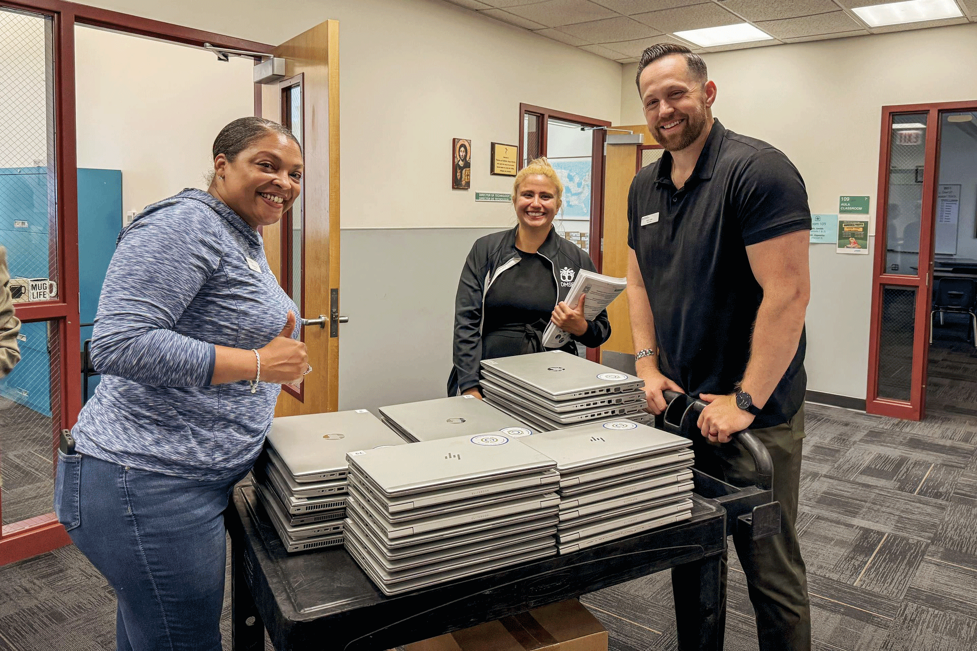 Two women and one man push a cart stacked with laptops