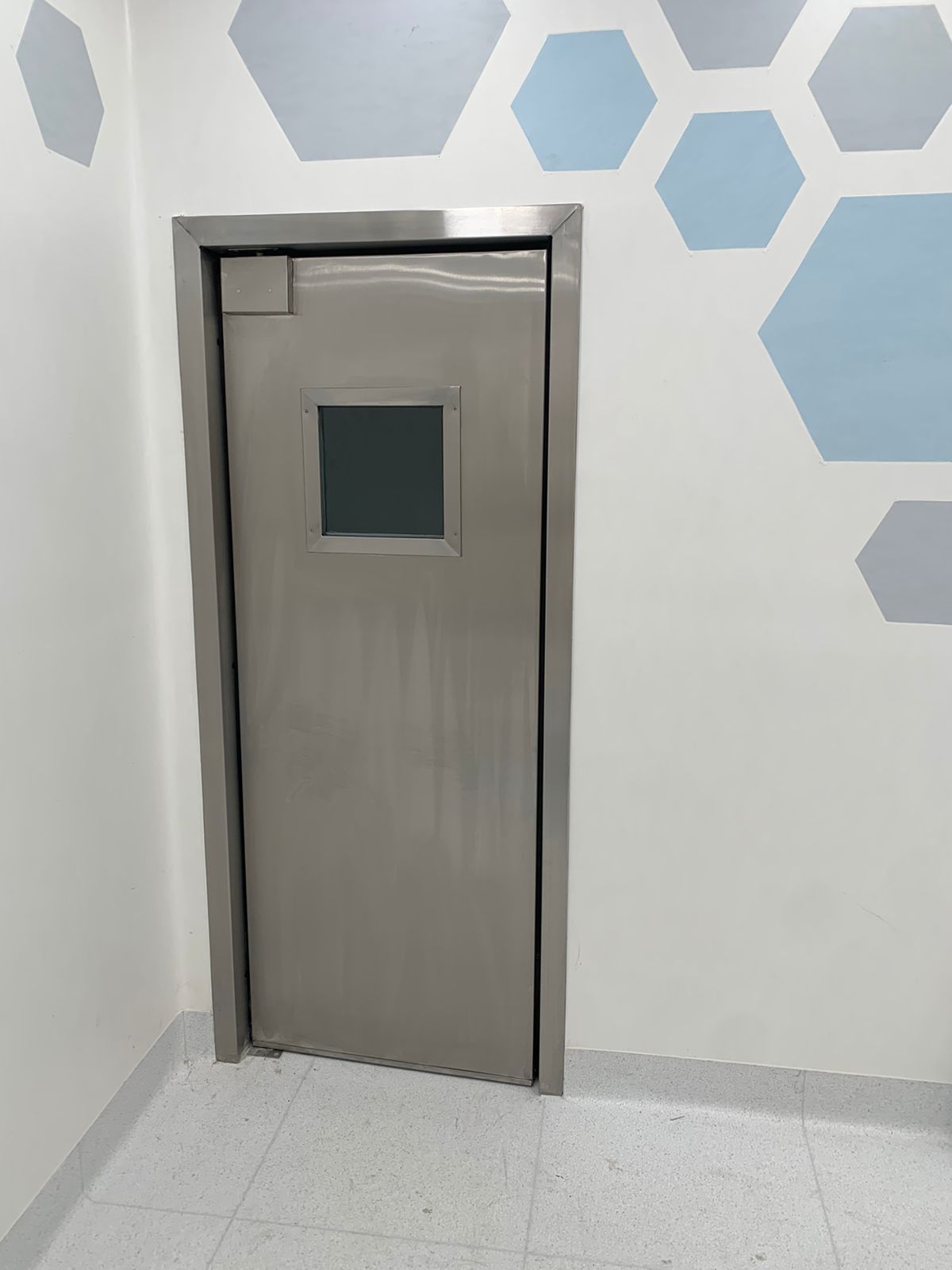 Una puerta de acero inoxidable con ventana en una habitación con hexágonos en la pared.