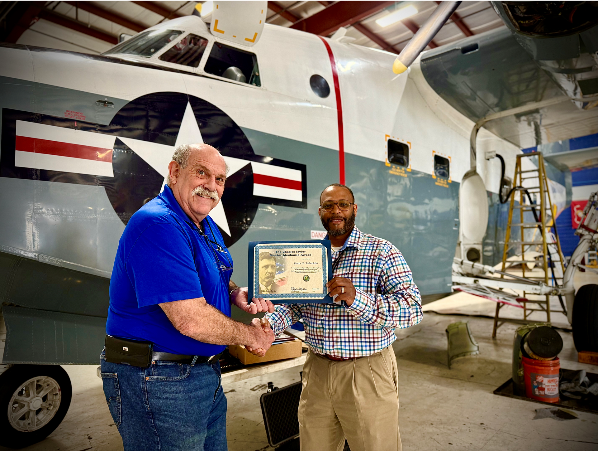Two men shaking hands in front of a seaplane, one holding an award.