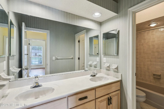 Bathroom with dual sinks, large mirror, and open doorway to a bathtub/shower. Light-colored cabinets and walls.