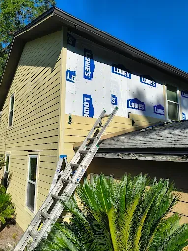 House exterior during siding replacement; ladder leaning against the wall; Lowe's house wrap exposed; sunny day.