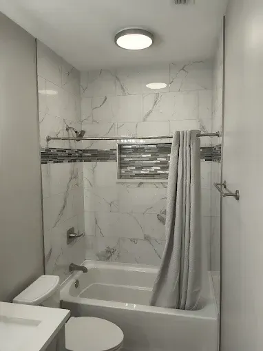 Bathroom with white marbled tiles, tub, toilet, gray shower curtain, and a recessed shelf.