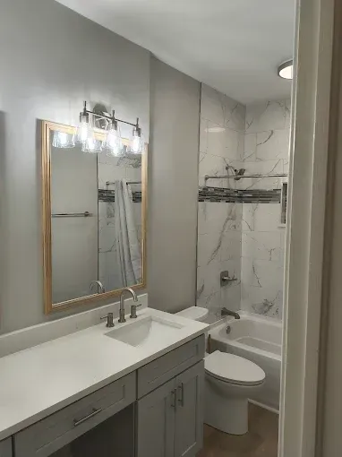Bathroom with light gray walls, vanity, mirror, toilet, and a bathtub with a marble tile surround.