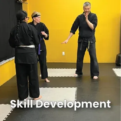 Three martial artists in black uniforms practice in a dojo with yellow walls. One demonstrates a move.