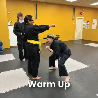Martial arts class: Instructor demonstrating a punch. Two students watch. Gym setting, yellow walls, gray mats.