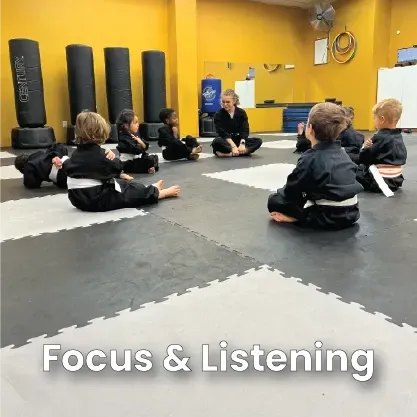 Children in black karate uniforms sit in a circle with an instructor on a mat. Yellow wall.