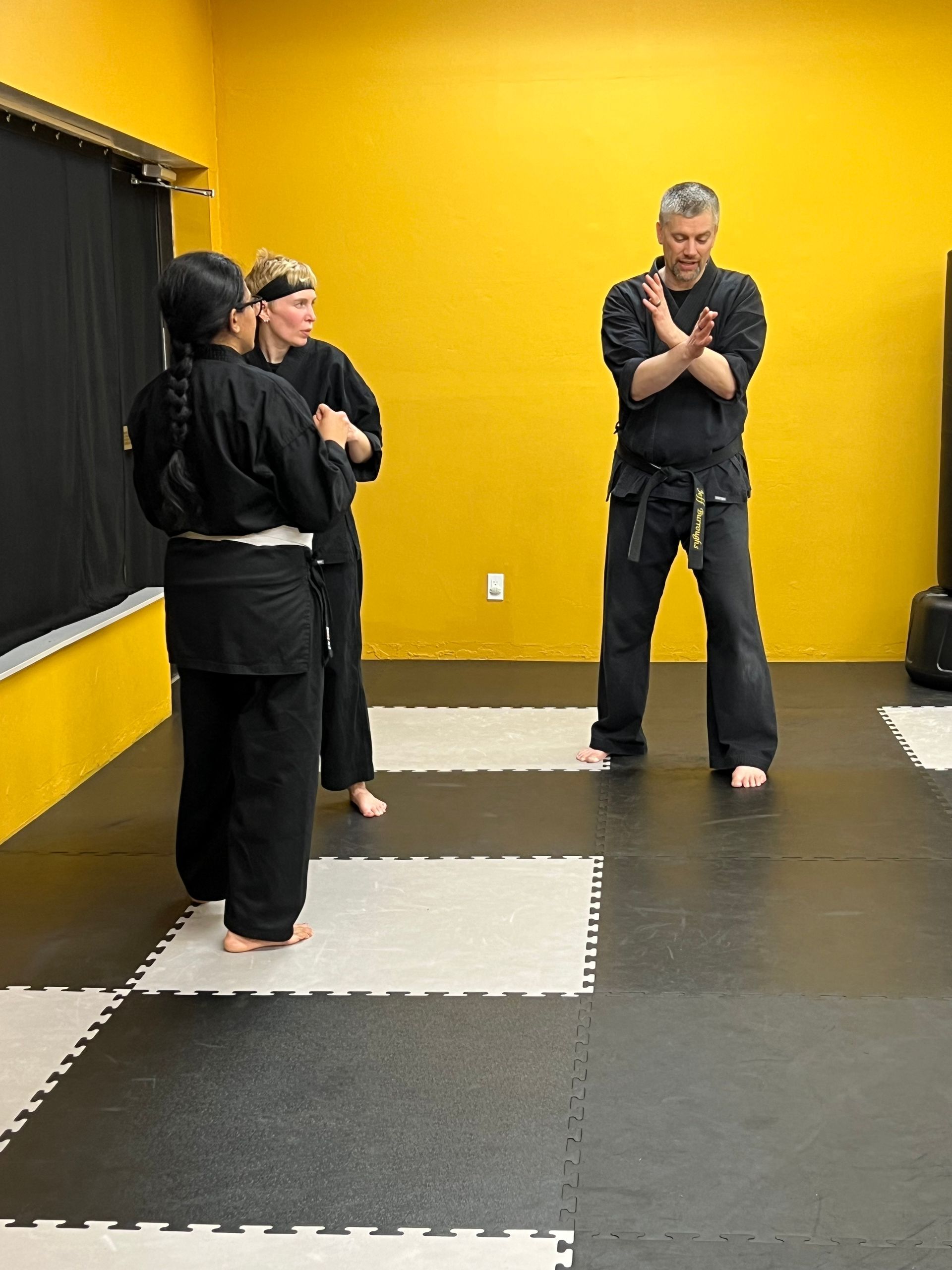Martial arts class: Three people in black uniforms on a mat, one demonstrating a move. Yellow wall in the background.