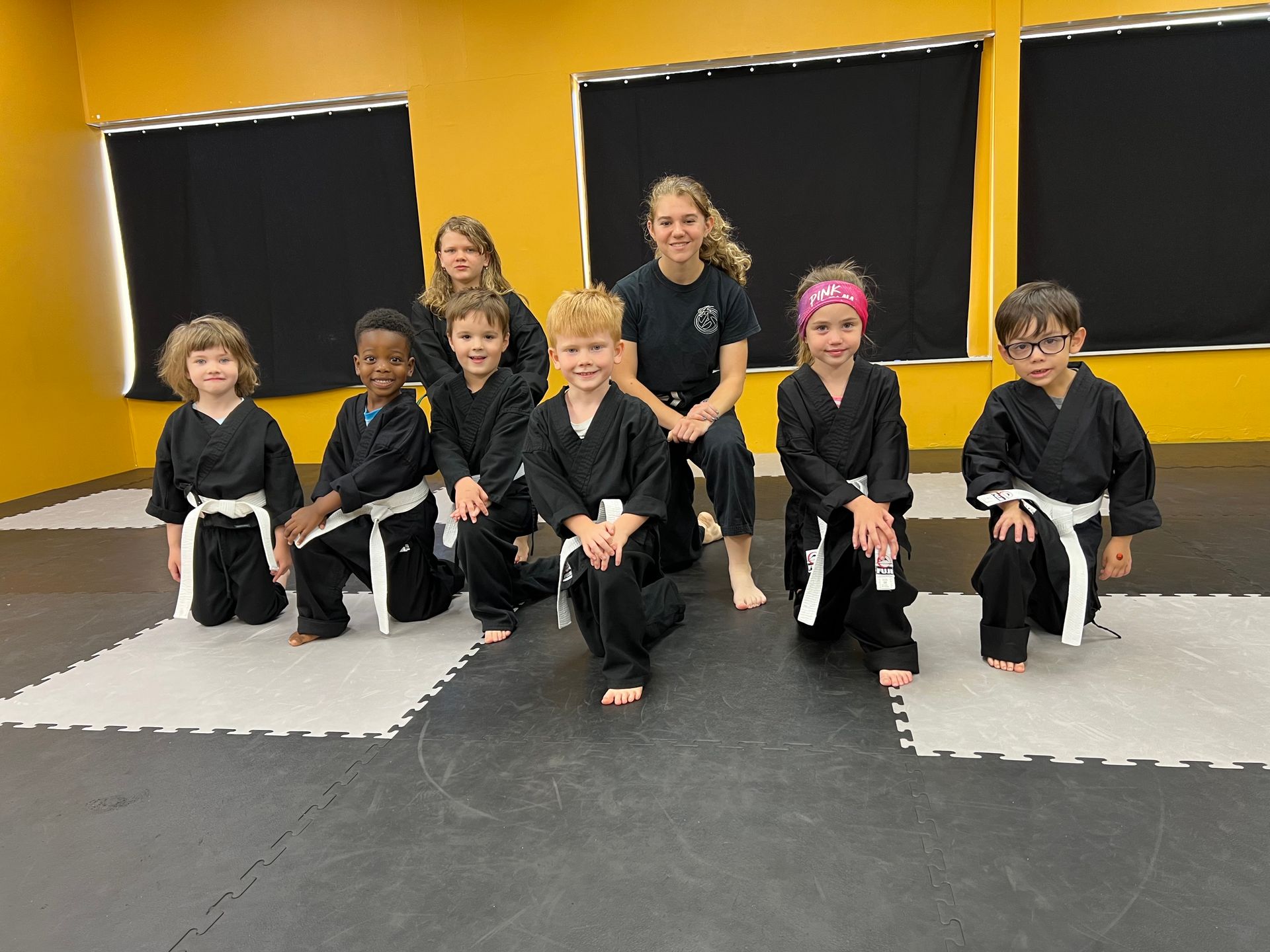 Group of children in black martial arts uniforms, kneeling with a female instructor on a mat.