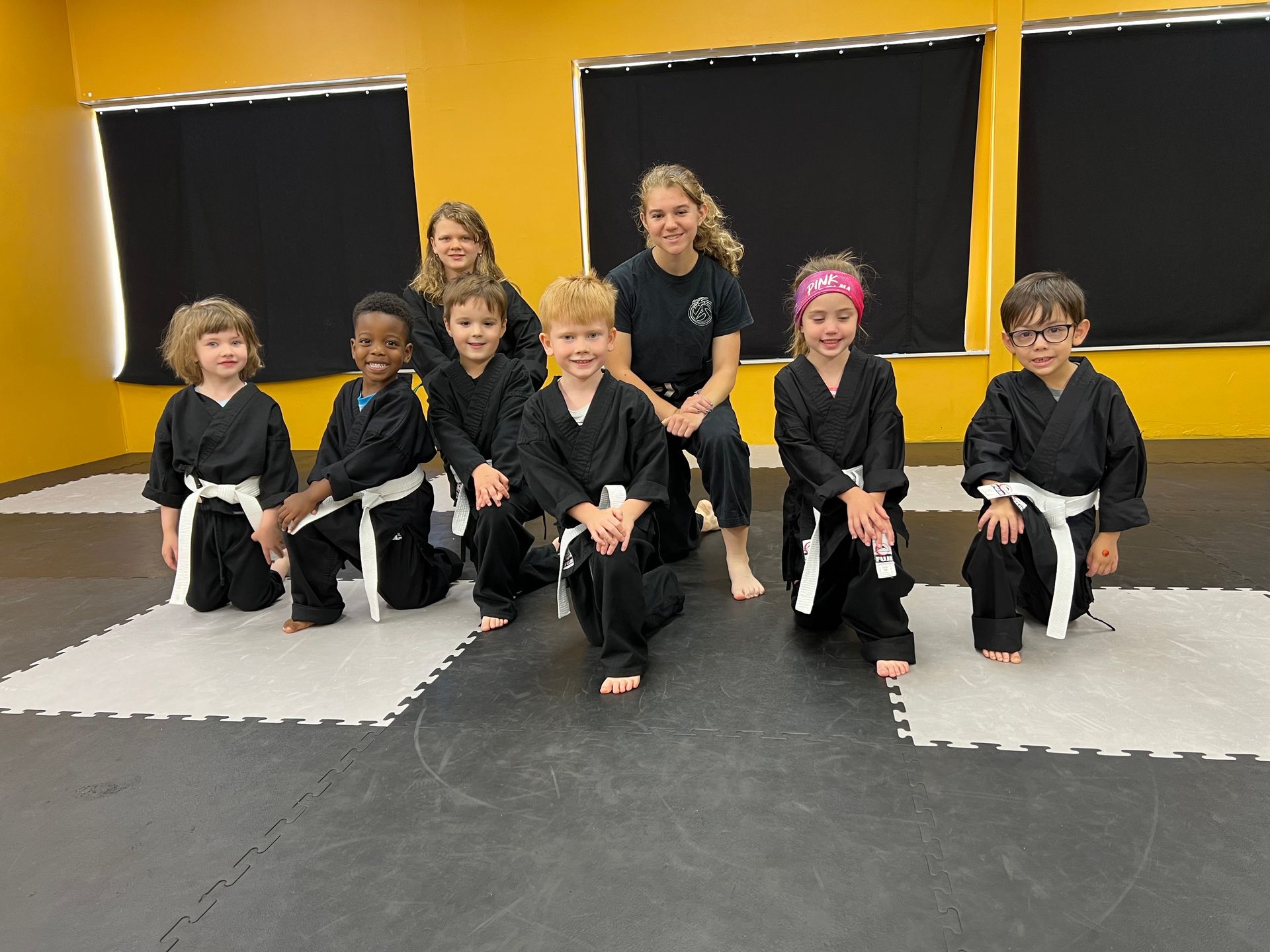 Group of children in black martial arts uniforms pose with two instructors in a dojo.