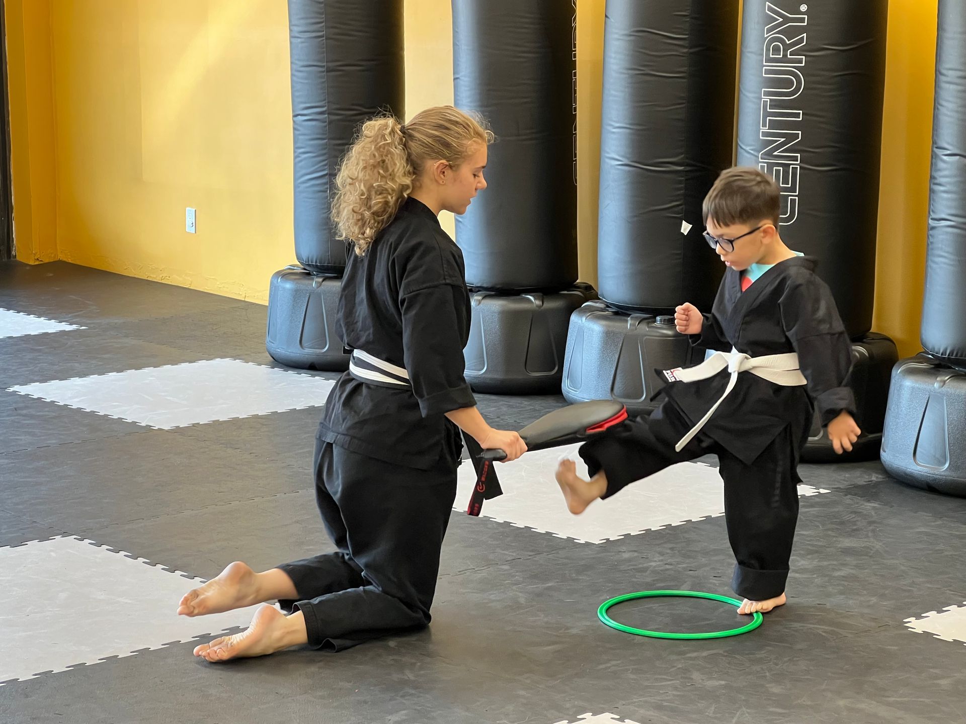 Woman in black uniform assisting child kicking a board in a martial arts class.
