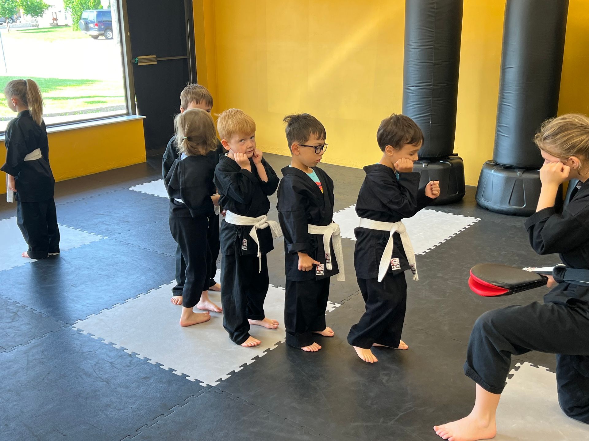 Children in black martial arts uniforms practicing a kick, instructed by someone in uniform, indoors.