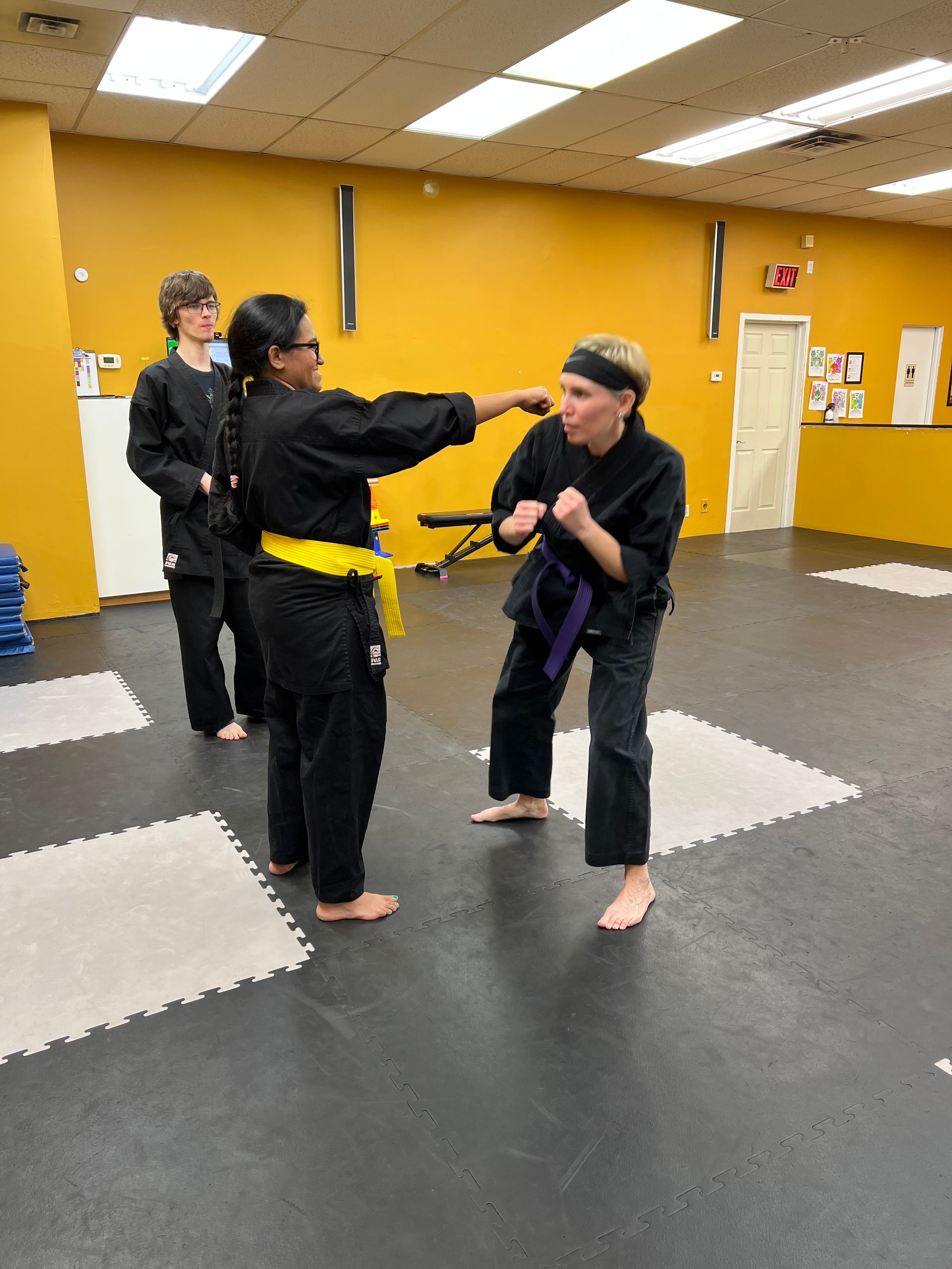 Martial arts class: a person in black uniform throws a punch at another person in purple belt uniform. Another student watches. Yellow and purple belts.