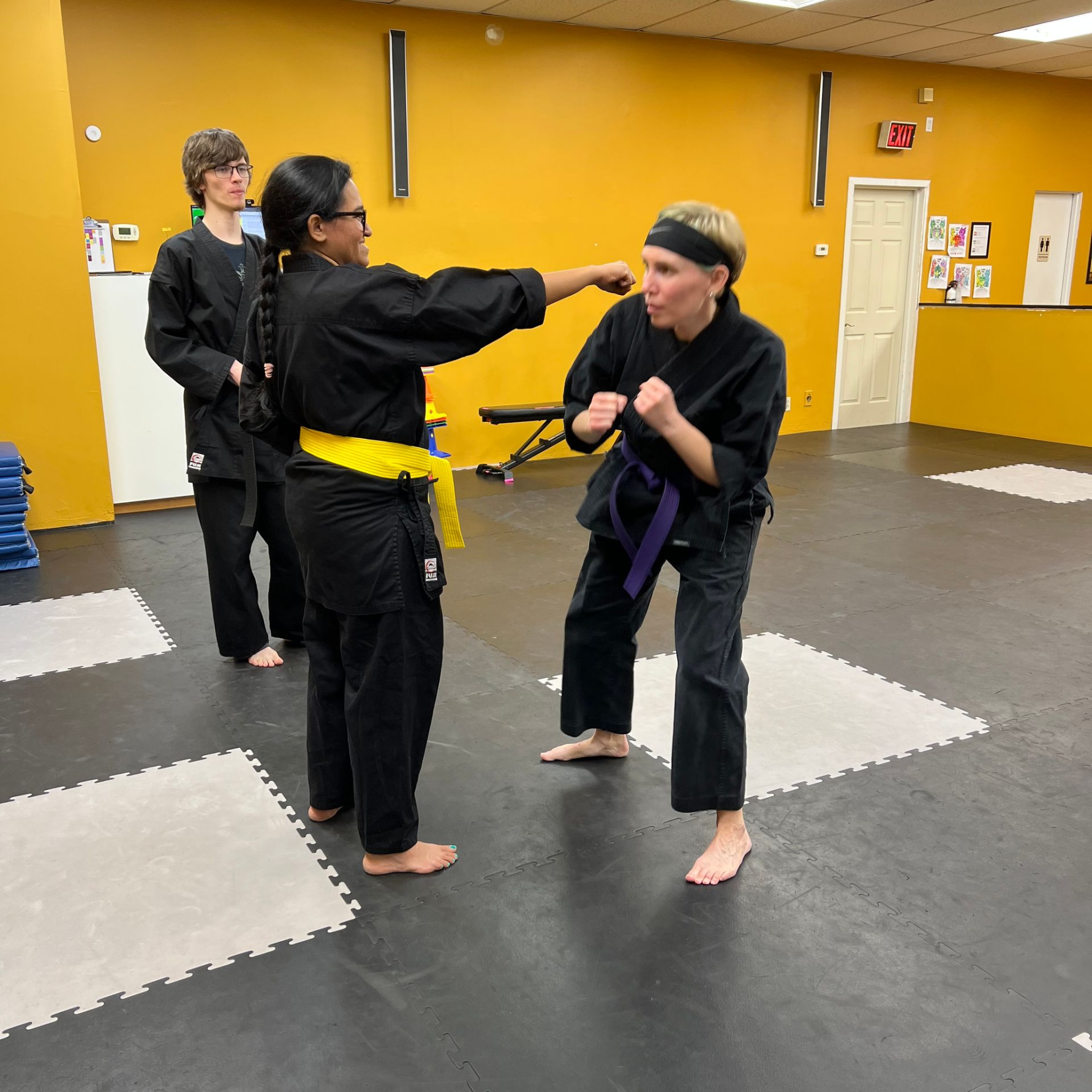 Martial arts class: Two students in black uniforms practice punches. Yellow and purple belts. Other student watches.