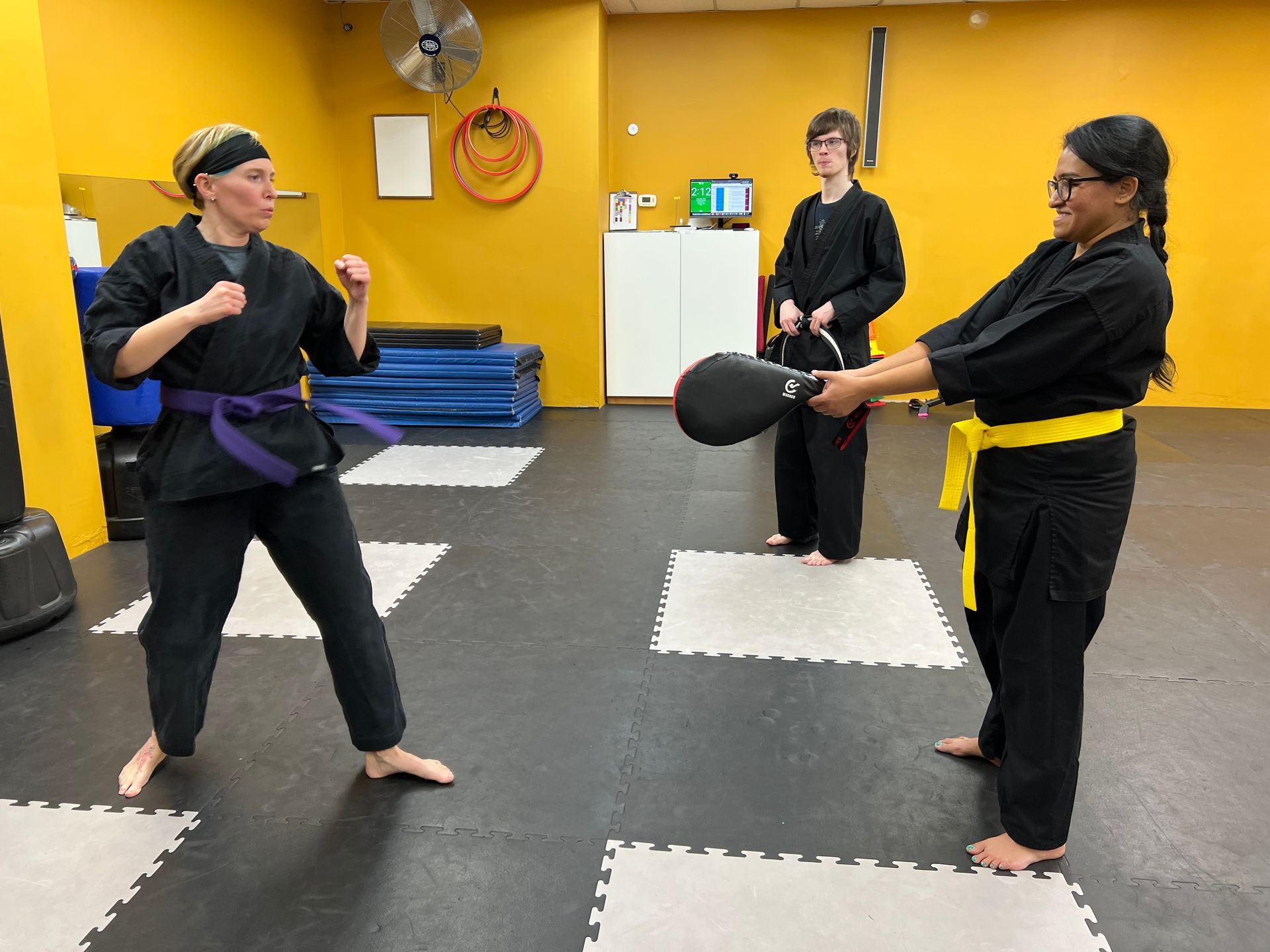 Martial arts class: Woman in black uniform kicking a target held by a person. Another person watches. Yellow walls.