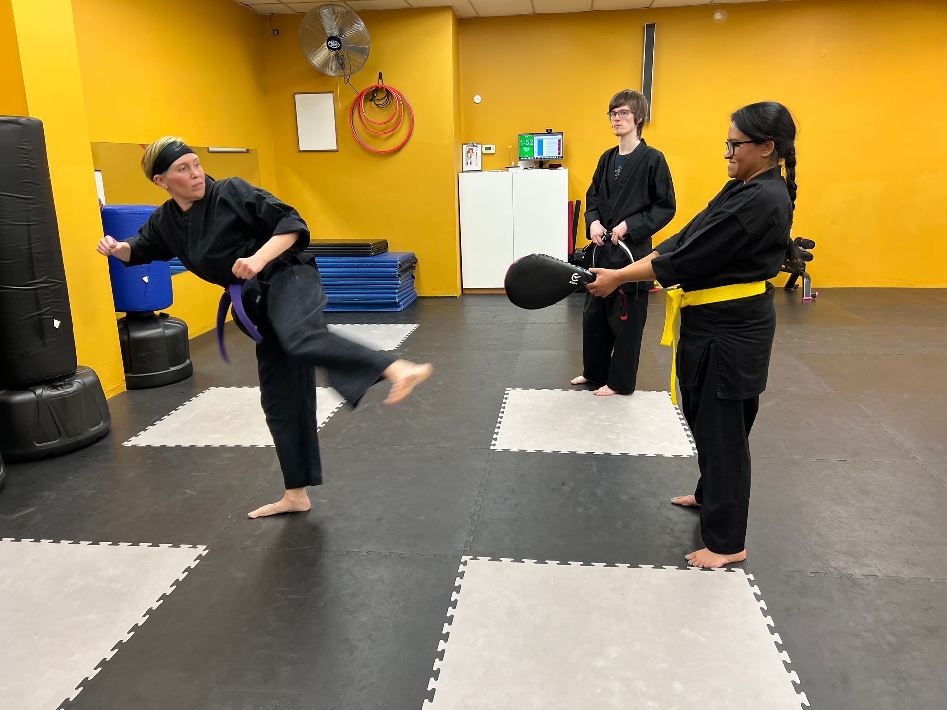 Martial arts class: Person kicks a bag held by another; two others watch in a dojo.