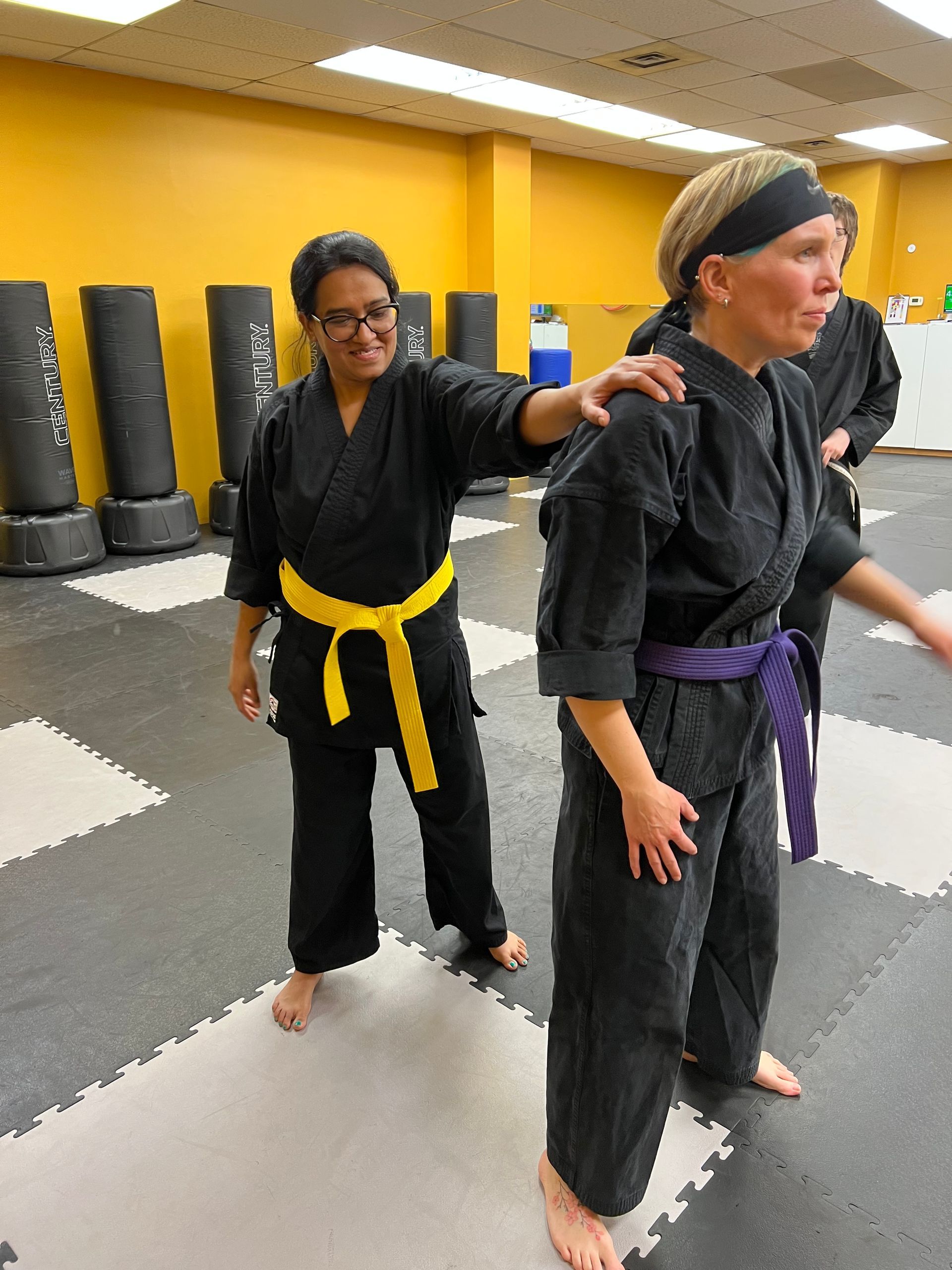 Two people in martial arts uniforms in a dojo. One with a yellow belt, smiles and touches the shoulder of the other with a purple belt.