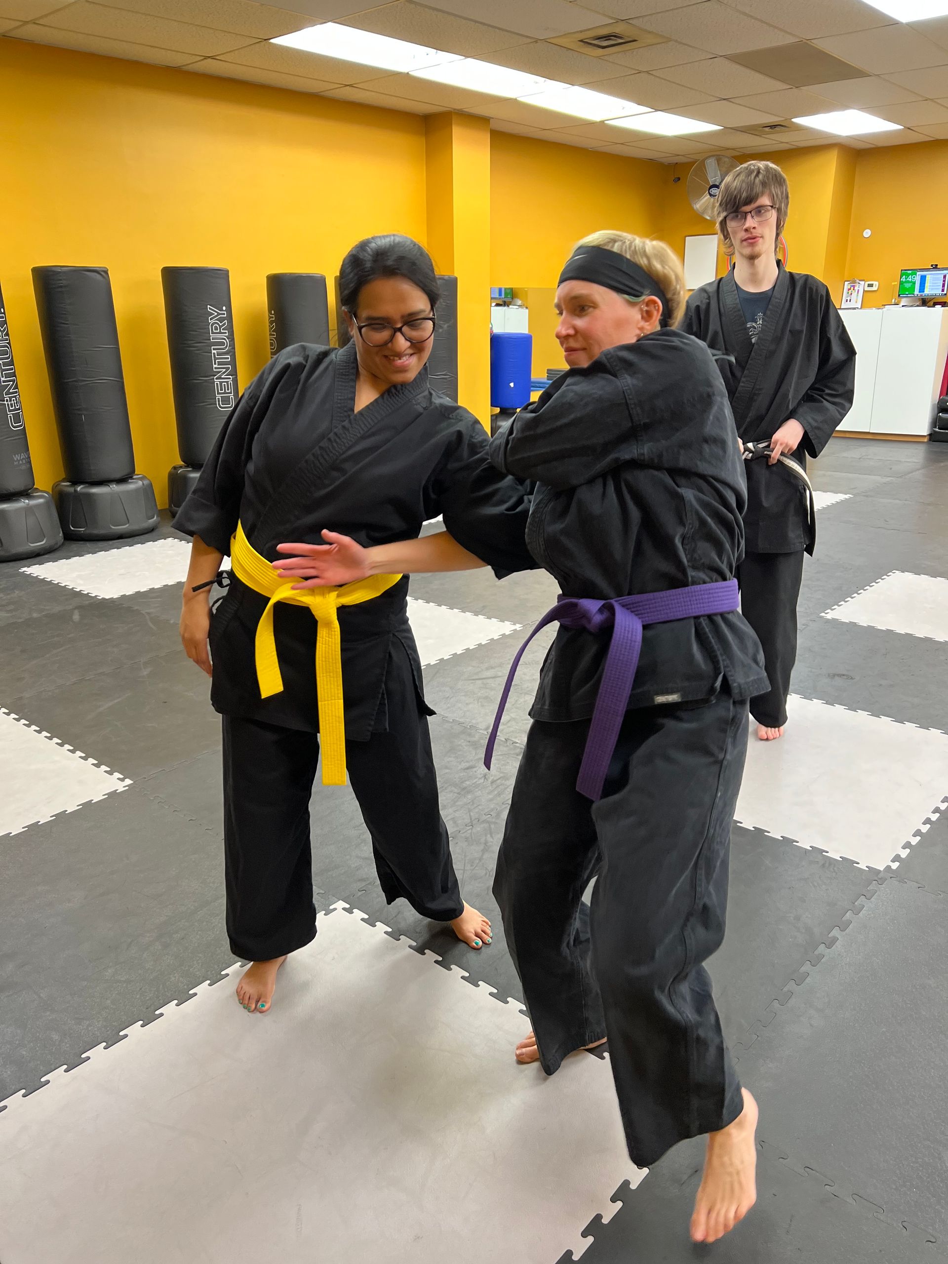 Two martial arts students in black uniforms practice a move in a dojo; one has a yellow belt, the other a purple belt.