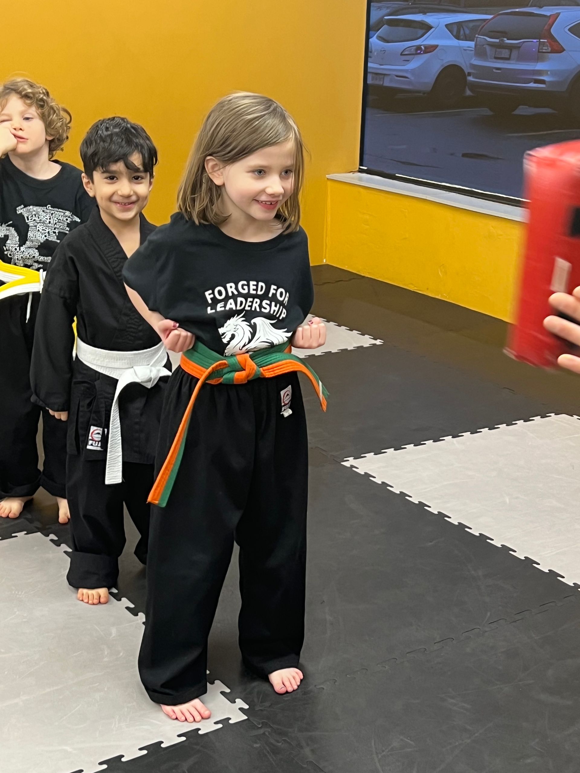 Children in karate uniforms, lined up in a dojo, preparing to strike a red pad.