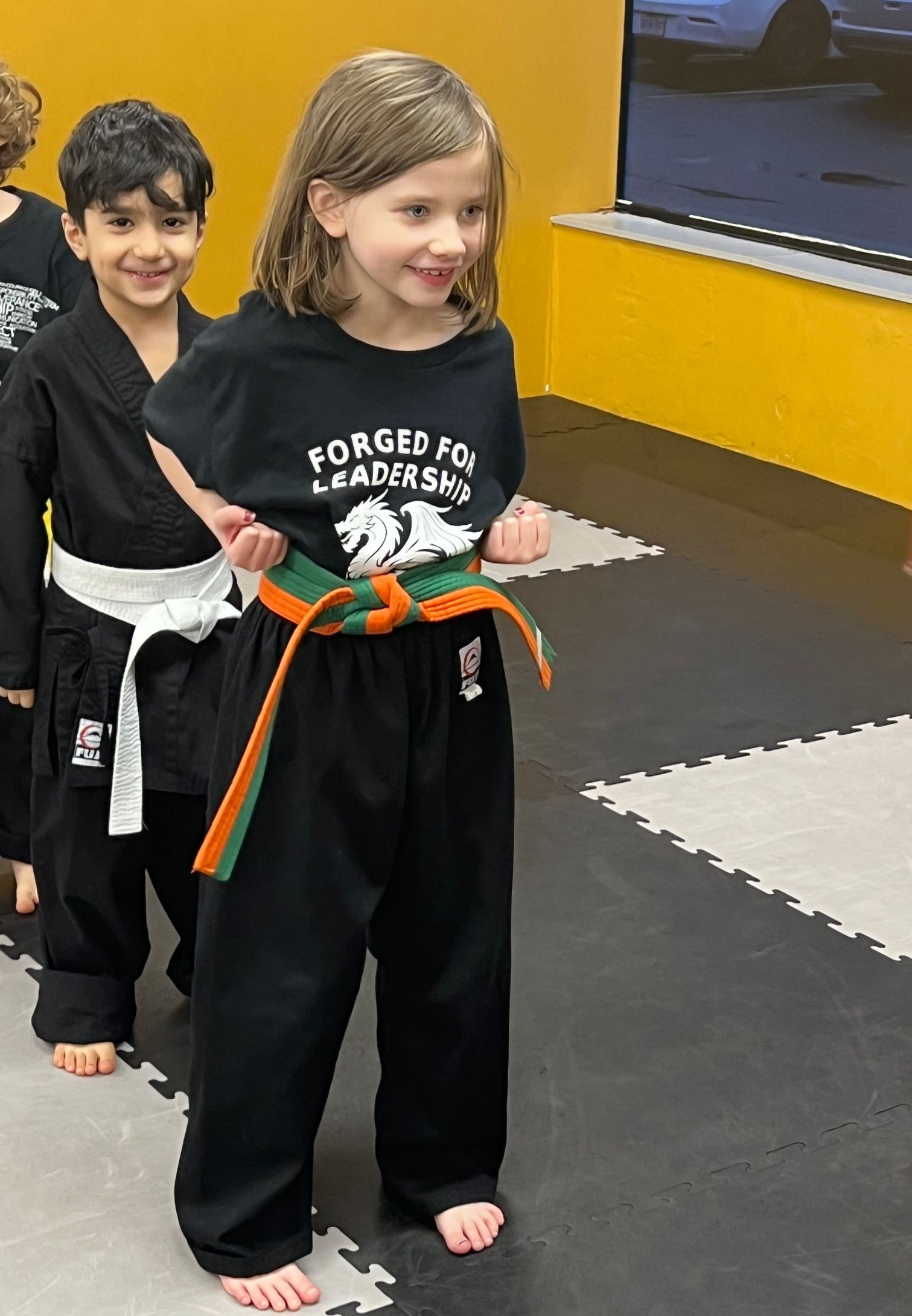 Girl in orange belt and black uniform smiles, standing with another child in a martial arts class.