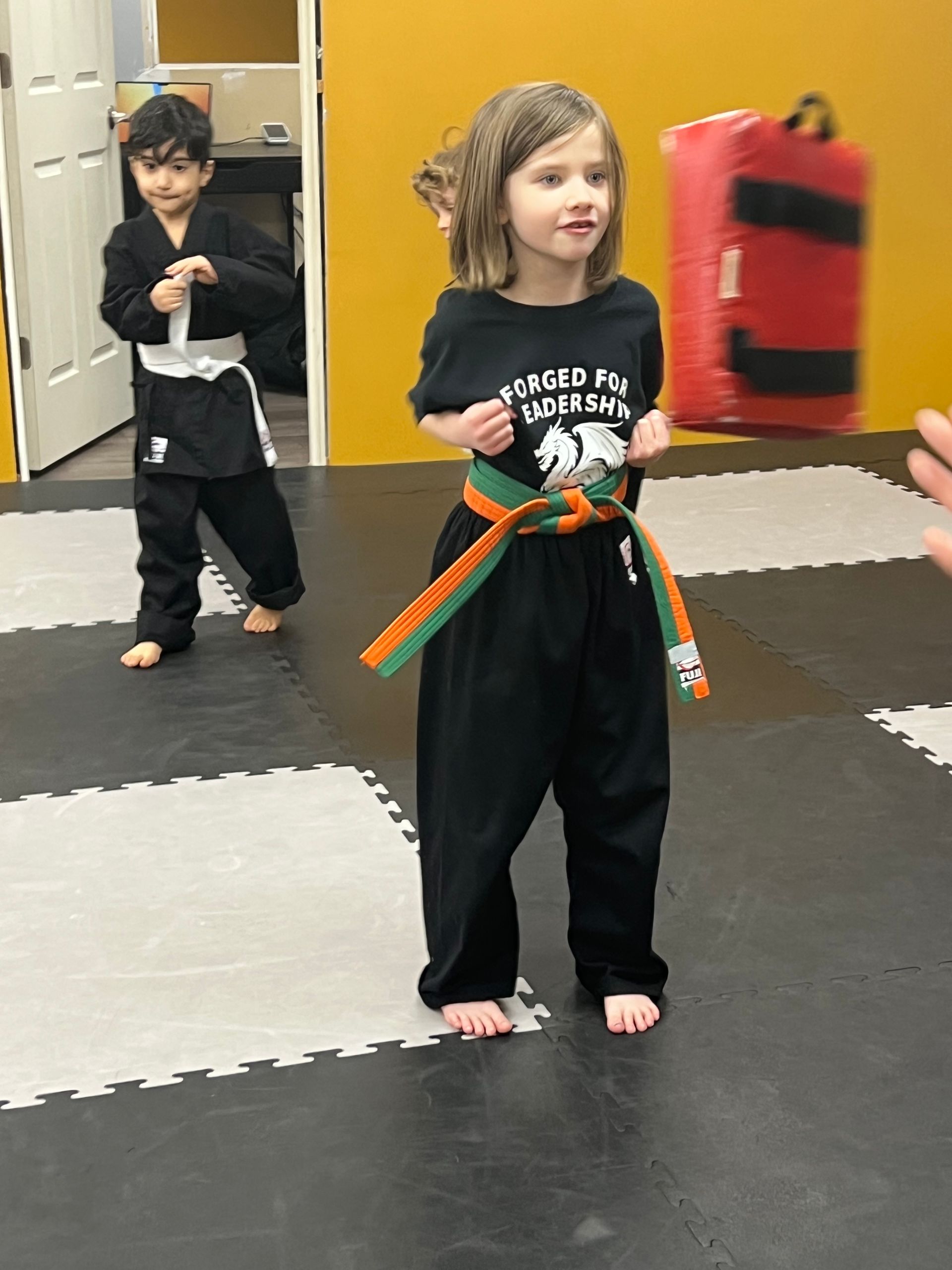 Two children in martial arts uniforms, one with an orange and green belt, inside a dojo.