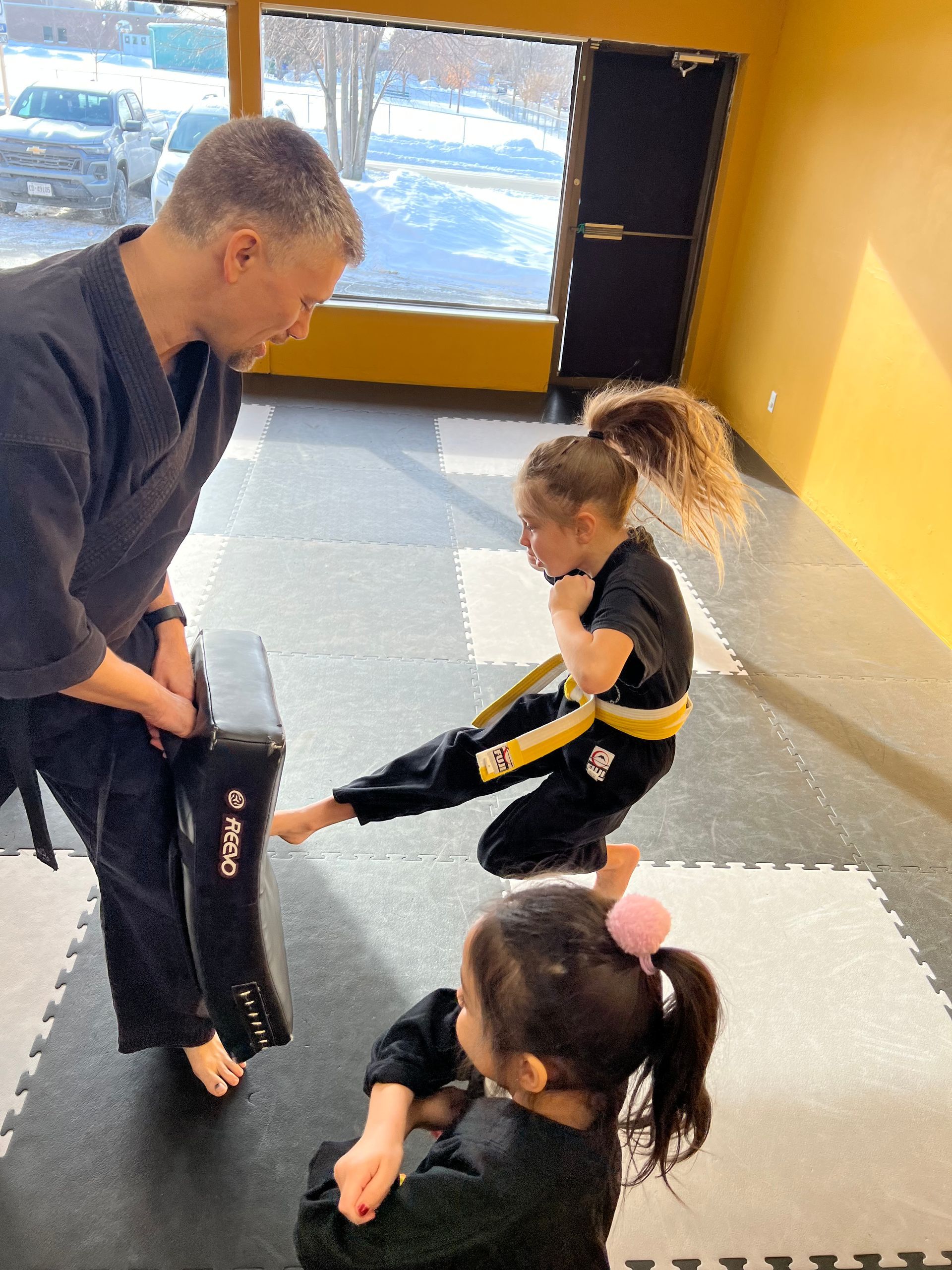Martial arts instructor assisting child in kicking exercise, in a dojo setting.