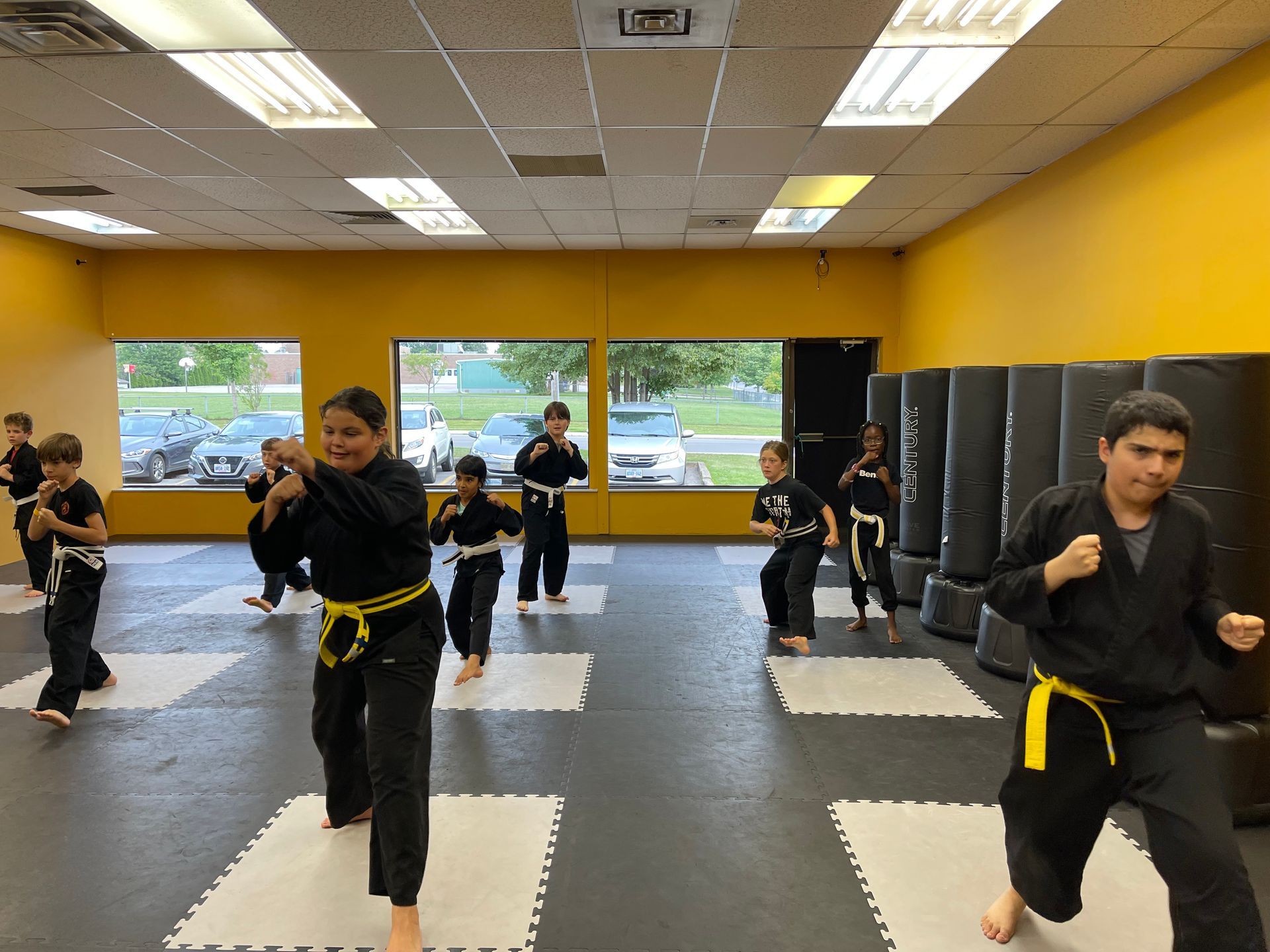 Group of children in black martial arts uniforms, kneeling with a female instructor on a mat.