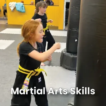 Boy in black martial arts uniform holding a striking pad, yellow belt. Training room.