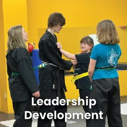 Children in black karate uniforms sit in a circle with an instructor on a mat. Yellow wall.