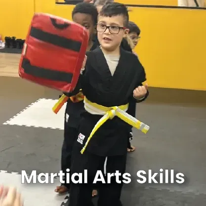 Boy in black martial arts uniform holding a striking pad, yellow belt. Training room.
