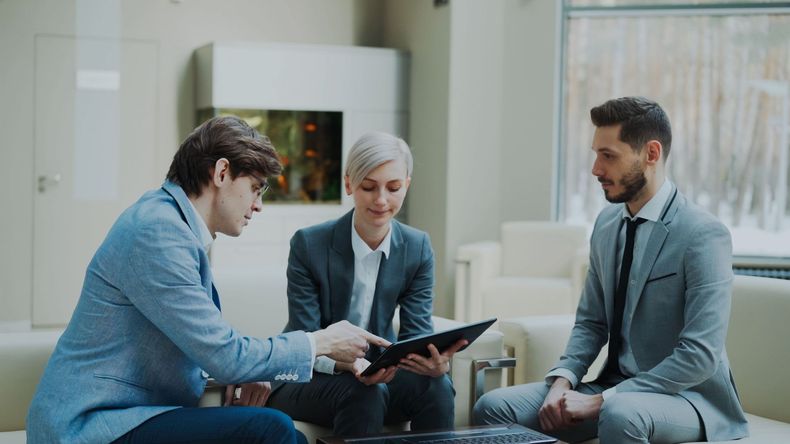 Three professionals in suits discuss documents in a modern office lounge.