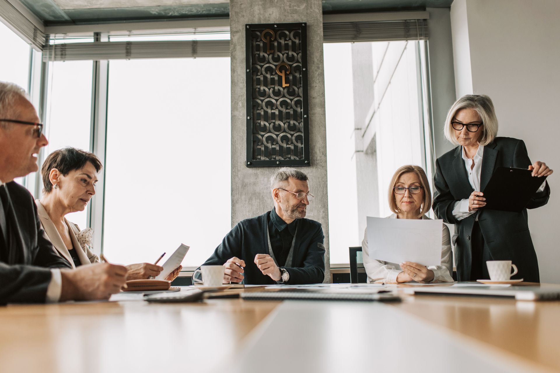 Business meeting in a bright conference room, with people seated around a table and one person standing and presenting.