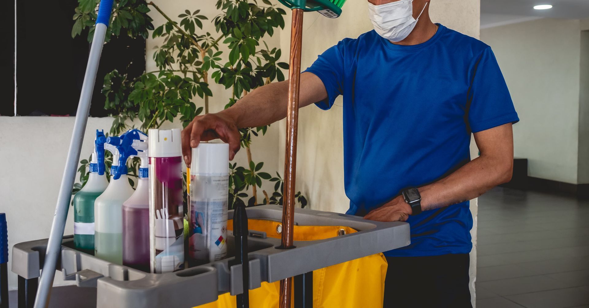 A person wearing a blue shirt and a face mask reaches for a cleaning spray on a cart filled with supplies and tools.