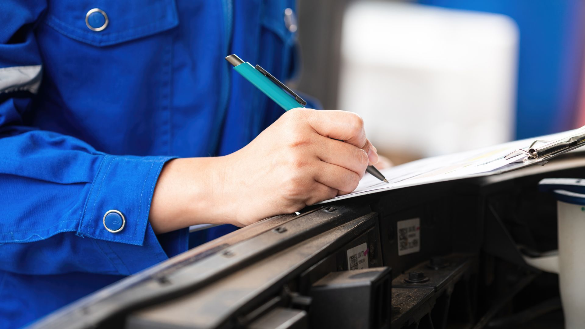 Mechanic in blue coveralls writing on a clipboard while inspecting a car engine.