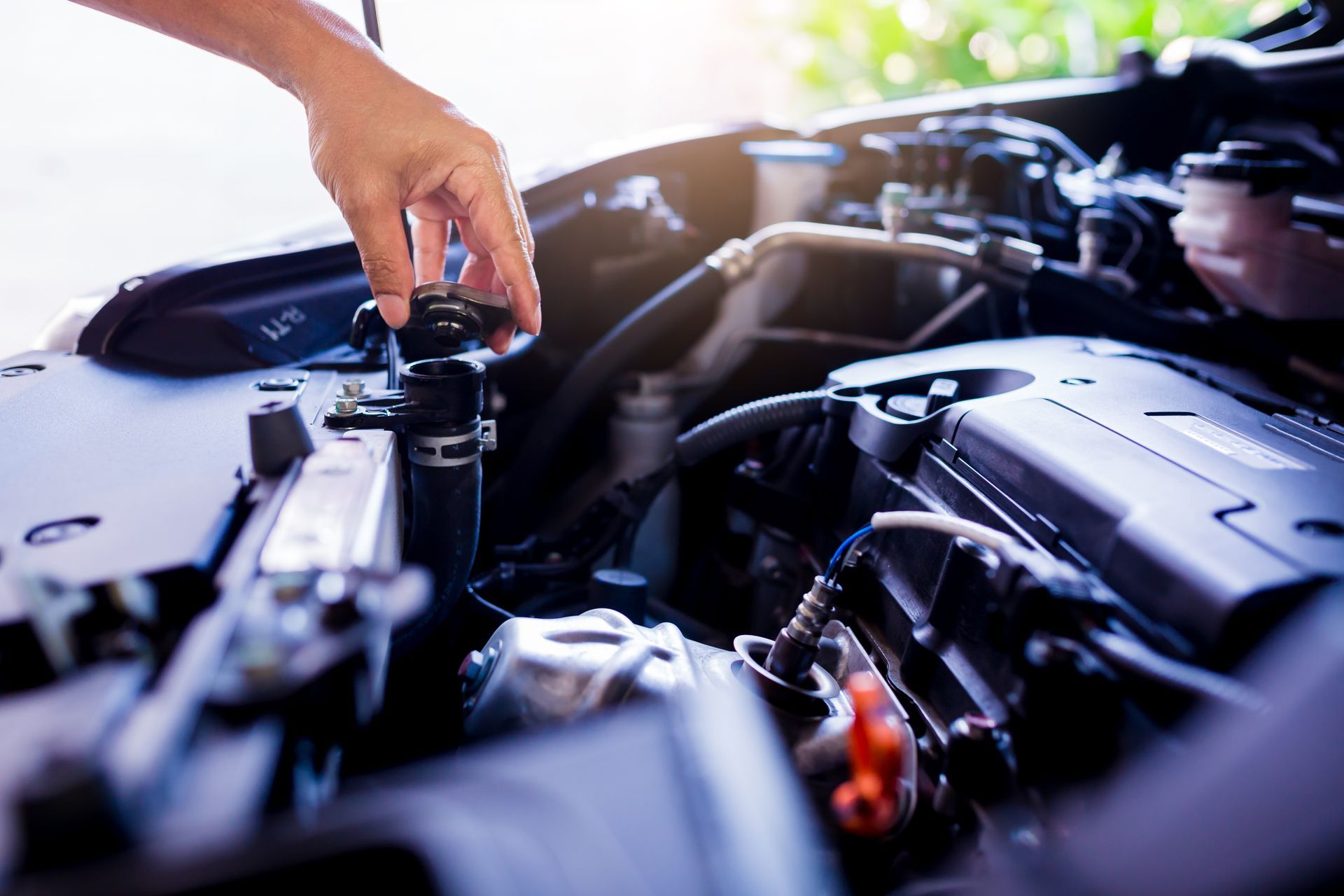 Person's hand opening radiator cap on a car engine. Bright outdoor setting.