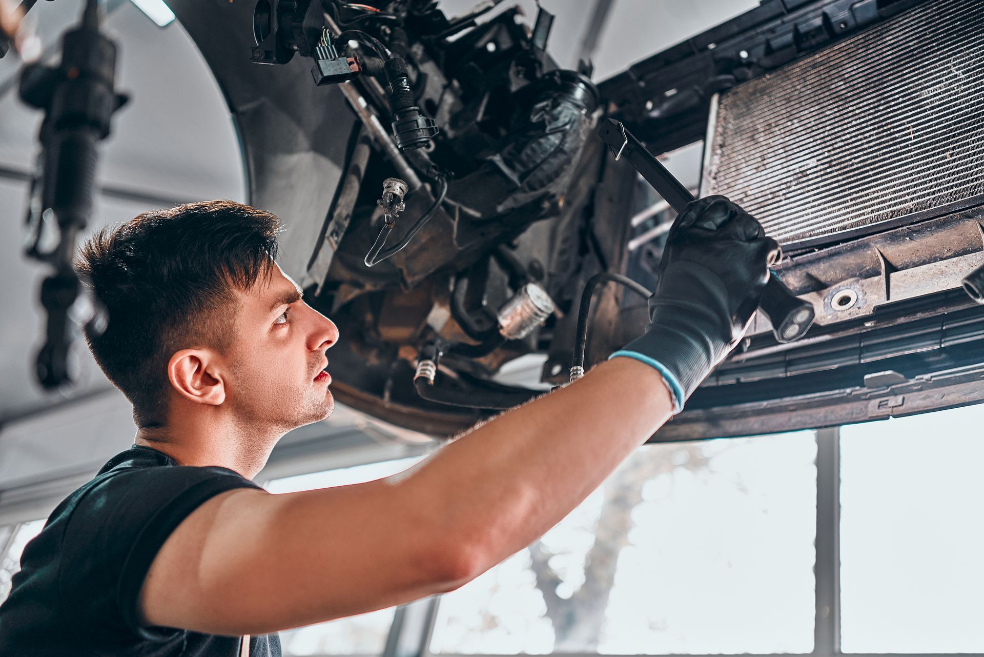 Mechanic working on the underside of a car, wearing gloves. Looking up with focused expression in a repair shop.