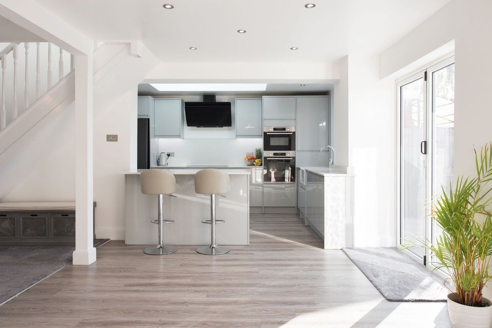 Open-plan kitchen with gray cabinets, bar stools, and sliding glass doors. Wooden floor, stairs on left, plant on right.
