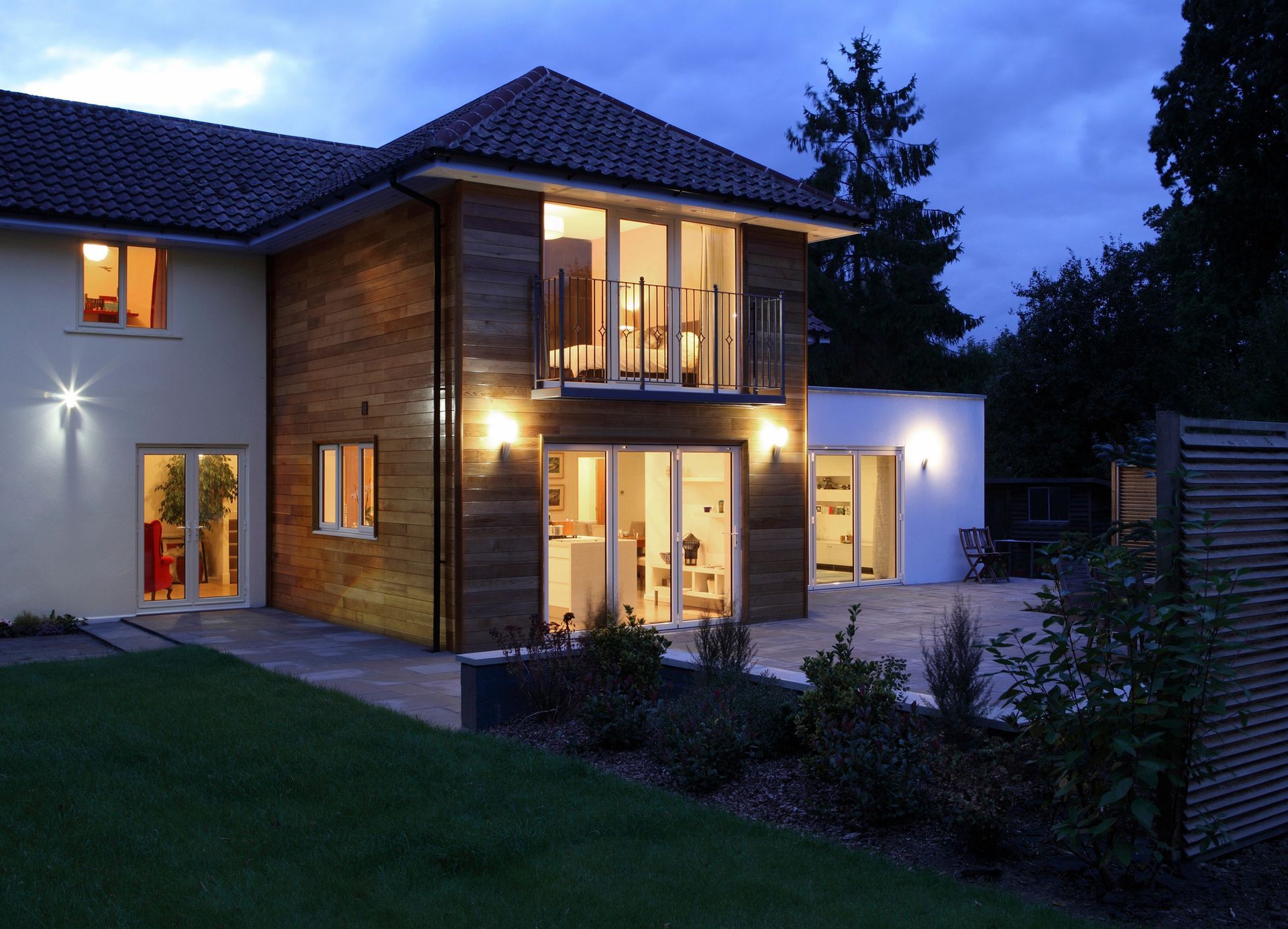 Two-story modern house at dusk, with lit windows, wood paneling, and a landscaped yard.