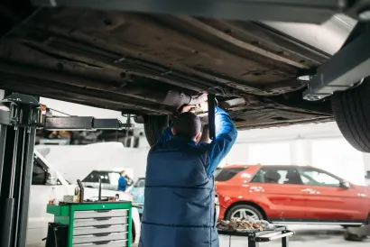 Mechanic working under a car raised on a lift in a repair shop; red SUV visible.