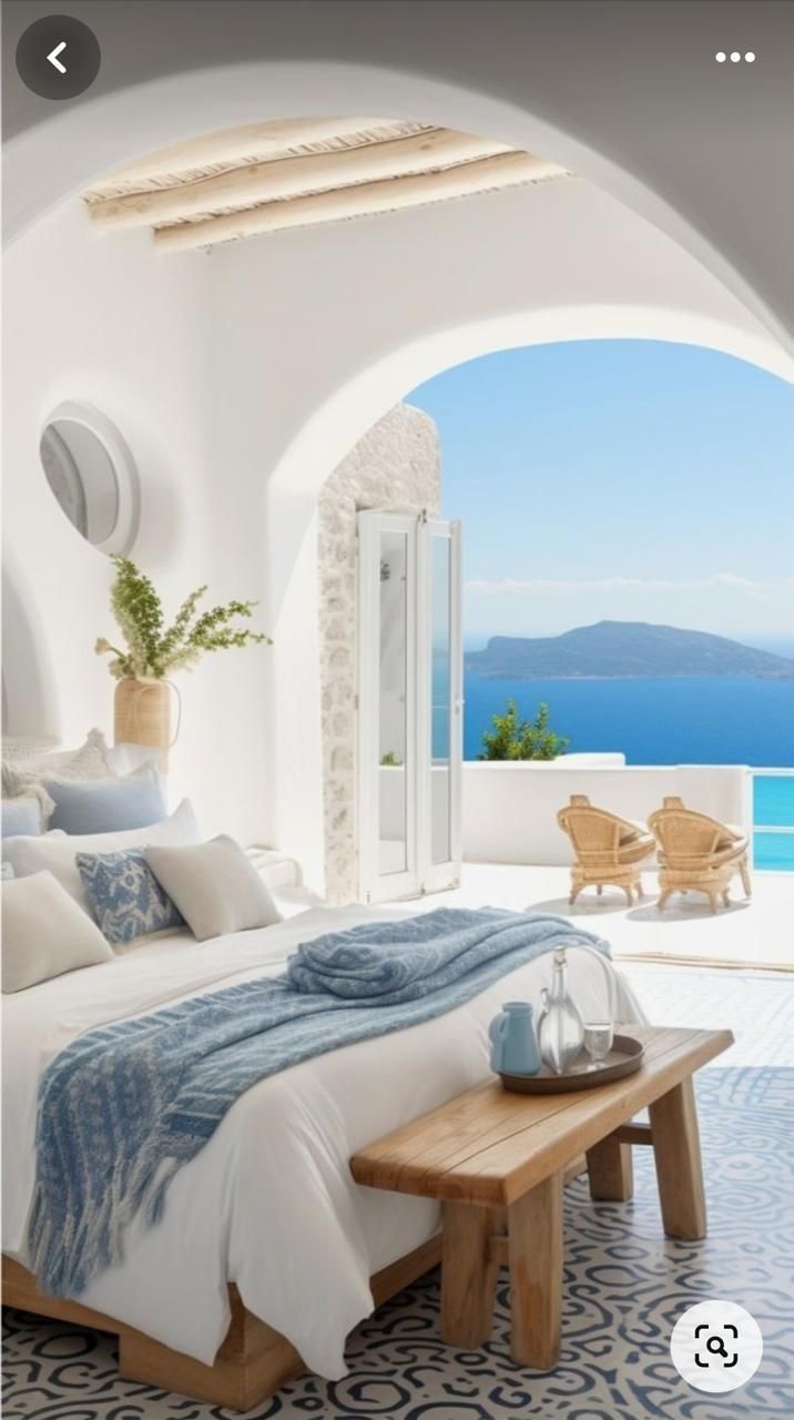 Dormitorio con puerta arqueada que da a un patio soleado con vistas al mar. Paredes blancas, detalles en azul y banco de madera.