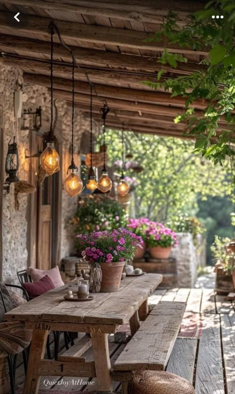 Comedor rústico al aire libre con mesa de madera, bancos, luces colgantes y flores en macetas.