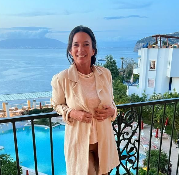 Una mujer con un blazer color canela se encuentra en un balcón, con el océano y la piscina al fondo.