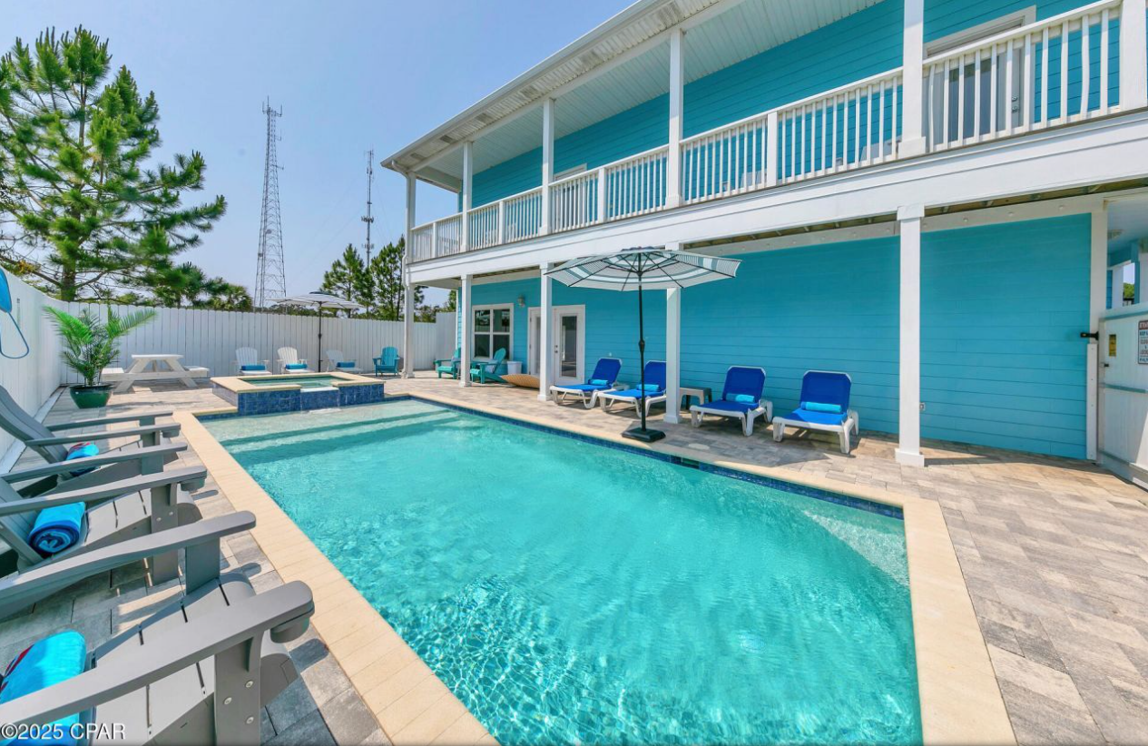 A vibrant blue, two-story house with a balcony overlooking a rectangular, sunlit swimming pool and patio area.