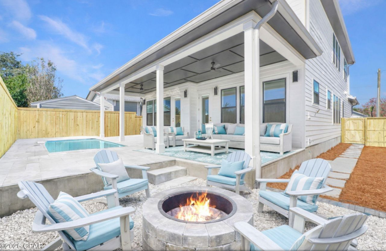 A sunny backyard patio with a pool, a fire pit, Adirondack chairs, and a covered porch with outdoor furniture.